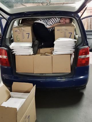 Eine Person liegt in einem Kofferraum eines blauen Autos, umgeben von Kartons und Paketen. Der Kofferraum ist voll mit Pappkartons und gestapelten Umschlägen. (Text KI generiert)