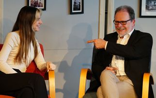 Zwei Personen sitzen in einem Gespräch auf einer Bühne. Die Frau mit langen Haaren lächelt, während der Mann in einem Anzug mit Brille sie anlächelt und mit dem Finger auf sie zeigt. Im Hintergrund sind Bilder an der Wand zu sehen. (Text KI generiert)