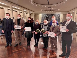 Eine Gruppe von sieben Personen steht in einem Raum, alle tragen Masken und halten Schilder mit Aufschriften. Einige Personen halten Blumen. Der Raum hat eine elegante Einrichtung mit Stühlen und einer dekorativen Wand im Hintergrund. (Text KI generiert)