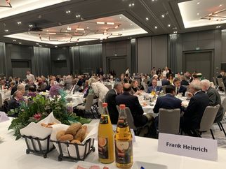 Ein elegantes Bankett mit vielen Gästen, die an runden Tischen sitzen und essen. Auf den Tischen stehen Getränke und frische Brötchen. Blumenarrangements sind ebenfalls sichtbar. Im Hintergrund sind weitere Gäste und eine moderne Raumgestaltung zu sehen. (Text KI generiert)