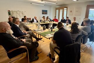 Eine Gruppe von etwa 15 Personen sitzt um einen Tisch in einem hellen Raum. Sie diskutieren und schauen auf Dokumente und Notizen. Im Hintergrund sind Fenster und eine Wand mit einem Kreuz und einem Bild. (Text KI generiert)