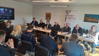 Eine Gruppe von Personen sitzt an einem Tisch in einem Konferenzraum. Einige Teilnehmer sind online zugeschaltet. Auf dem Tisch liegen Notizen und Laptops. Im Hintergrund sind Banner und Bilder sichtbar, die mit dem Thema Religionsfreiheit verbunden sind. (Text KI generiert)