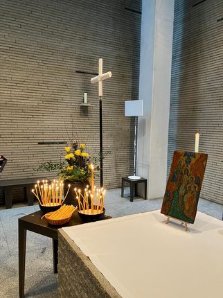 Ein Innenraum mit einem Tisch, auf dem eine Ikone steht. Daneben brennen Kerzen in einem Korb. Im Hintergrund sind Blumen und ein Kreuz sichtbar. Die Wände sind aus Stein, und es gibt eine Lampe. (Text KI generiert)
