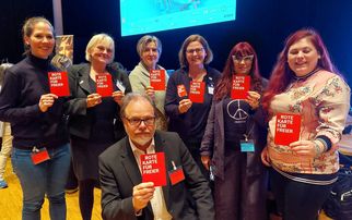 Eine Gruppe von acht Personen steht zusammen und hält rote Karten mit der Aufschrift "ROTE KARTE FÜR FREIER" in die Kamera. Sie lächeln und scheinen an einer Veranstaltung teilzunehmen. Im Hintergrund ist eine Leinwand sichtbar. (Text KI generiert)