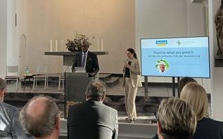 Eine Präsentation in einem modernen Kirchenraum. Im Vordergrund stehen zwei Personen am Rednerpult, während das Publikum auf Bänken sitzt. Im Hintergrund ist ein Bildschirm mit Informationen zu sehen. Der Raum ist hell erleuchtet und hat eine schlichte, elegante Gestaltung. (Text KI generiert)