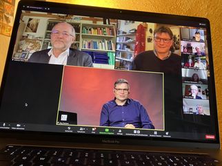 Eine Zoom-Konferenz auf einem Laptop-Bildschirm zeigt mehrere Teilnehmer in verschiedenen Fenstern. Im Vordergrund sitzt ein Mann mit Brille und dunkler Kleidung, während im Hintergrund zwei weitere Männer und mehrere weitere Teilnehmer in kleineren Fenstern zu sehen sind. (Text KI generiert)