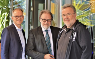 Drei Männer stehen vor einer Glastür, umgeben von Pflanzen. Der Mann links trägt eine Brille und einen Anzug, der Mann in der Mitte hat graue Haare und einen Anzug mit Krawatte, der Mann rechts trägt ein schwarzes T-Shirt und eine Jacke. Alle lächeln. (Text KI generiert)