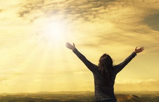 Eine Person steht mit ausgebreiteten Armen auf einem Hügel und blickt in den Himmel. Die Sonne strahlt hell und der Himmel ist in warmen Gelbtönen gefärbt. Im Hintergrund sind sanfte Hügel und eine weite Landschaft zu sehen. (Text KI generiert)