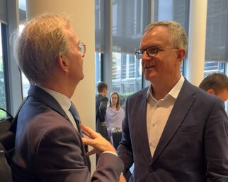 Zwei Männer stehen in einem modernen Raum und unterhalten sich freundlich. Der Mann links trägt einen Anzug und eine Krawatte, während der Mann rechts ein helles Hemd und einen dunklen Blazer trägt. Im Hintergrund sind weitere Personen zu sehen.