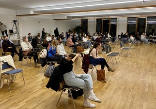 Eine Gruppe von Menschen sitzt in einem großen Raum auf Stühlen, mit Abstand zueinander. Einige Personen tragen Masken. Im Hintergrund sind Fenster und Pflanzen sichtbar. Der Raum hat einen hellen Holzfußboden und eine ruhige Atmosphäre. (Text KI generiert)