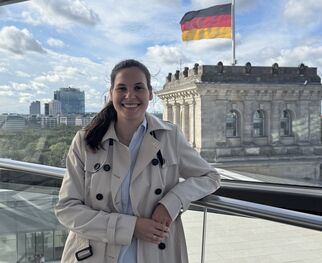 Eine Person in einem beigen Trenchcoat steht an einem Glasgeländer und lächelt. Im Hintergrund ist das Reichstagsgebäude mit der deutschen Flagge zu sehen. Der Himmel ist teilweise bewölkt. (Text KI generiert)