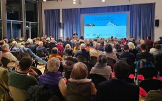 Eine große Gruppe von Menschen sitzt in einem Raum und hört einem Vortrag zu. Im Hintergrund ist eine Leinwand mit Präsentationstext und Bildern zu sehen. Die Atmosphäre wirkt informativ und aufmerksam. (Text KI generiert)