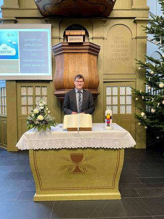 Ein Mann steht an einem Altar in einer Kirche, umgeben von Blumen und Kerzen. Im Hintergrund ist eine Präsentation zu sehen. Der Altar ist aus Holz und hat eine goldene Farbe. Rechts steht ein Weihnachtsbaum. (Text KI generiert)