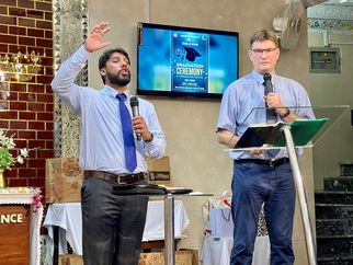 Zwei Männer stehen auf einer Bühne während einer Abschlusszeremonie. Der Mann links spricht mit erhobener Hand, während der Mann rechts ein Mikrofon hält und in ein Notizbuch schaut. Im Hintergrund ist ein Bildschirm mit Informationen zur Zeremonie zu sehen. (Text KI generiert)