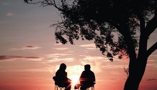 Zwei Personen sitzen in Stühlen unter einem Baum und beobachten den Sonnenuntergang. Die Silhouetten sind vor einem bunten Himmel sichtbar, der von warmen Farben geprägt ist. (Text KI generiert)