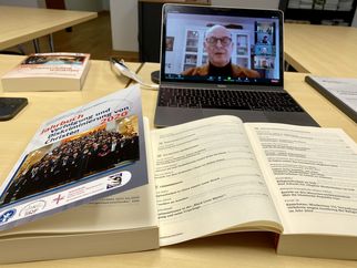 Auf einem Tisch liegen ein offenes Buch und ein Jahrbuch mit dem Titel "Jahrbuch und Verzeichnis von Christen 2020". Im Hintergrund ist ein Laptop mit einer Videoanrufansicht zu sehen, auf dem eine Person spricht. (Text KI generiert)